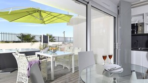 A modern outdoor dining area with a glass table, white wicker chairs, and a green umbrella. - NAUTILIA  103 - Premium Luxe - Jacuzzi - 2 people (AGDE)