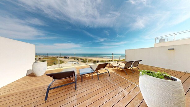 Una terraza de madera con sillones de descanso y una jardinera, con vista a una playa y el océano.