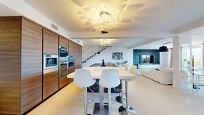 Una cocina moderna con electrodomésticos integrados, un comedor con sillas blancas, una escalera y una sala de estar con televisor.