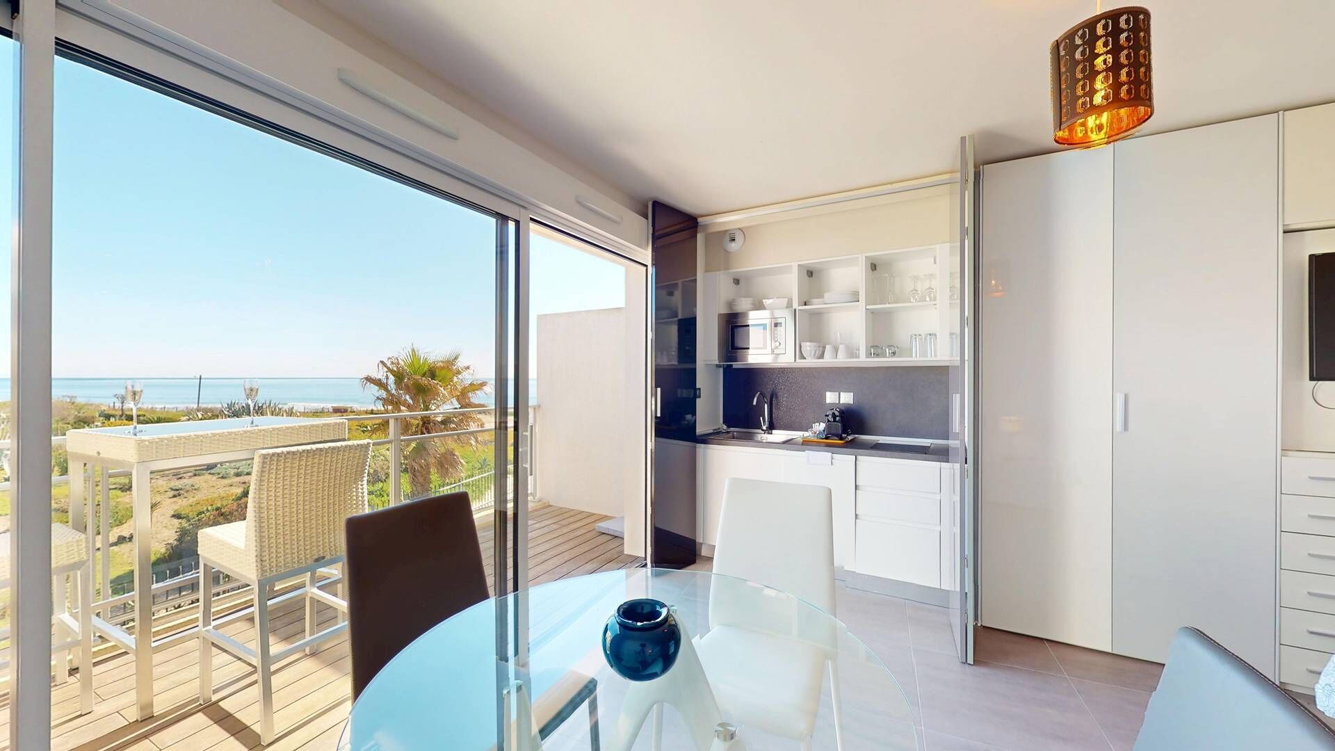 A balcony with a view of the sea, a glass table with a vase, and a kitchen with white cabinets.