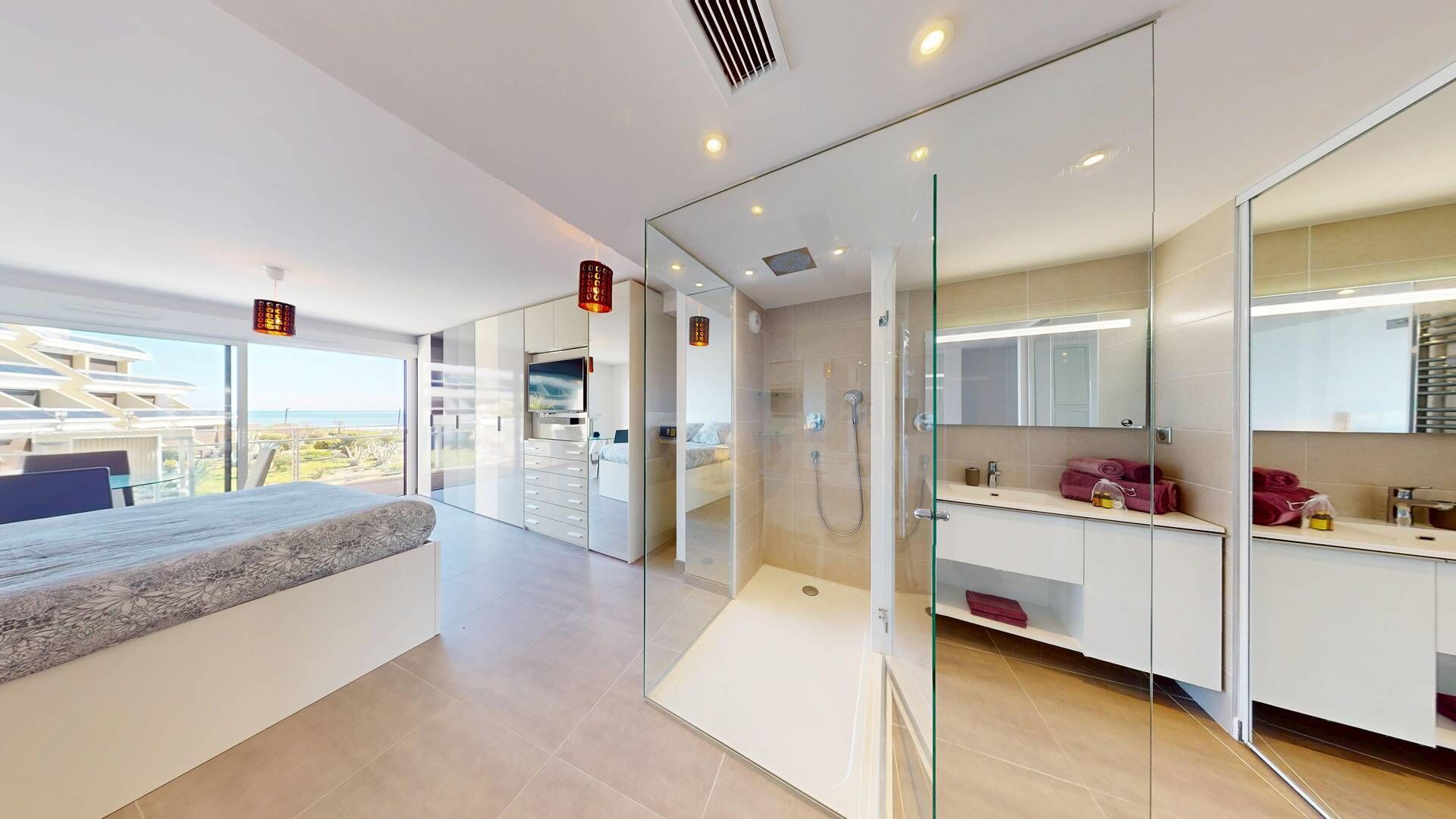 A modern hotel room with a bed, a glass-enclosed shower, a vanity with a mirror, and a view of the ocean.