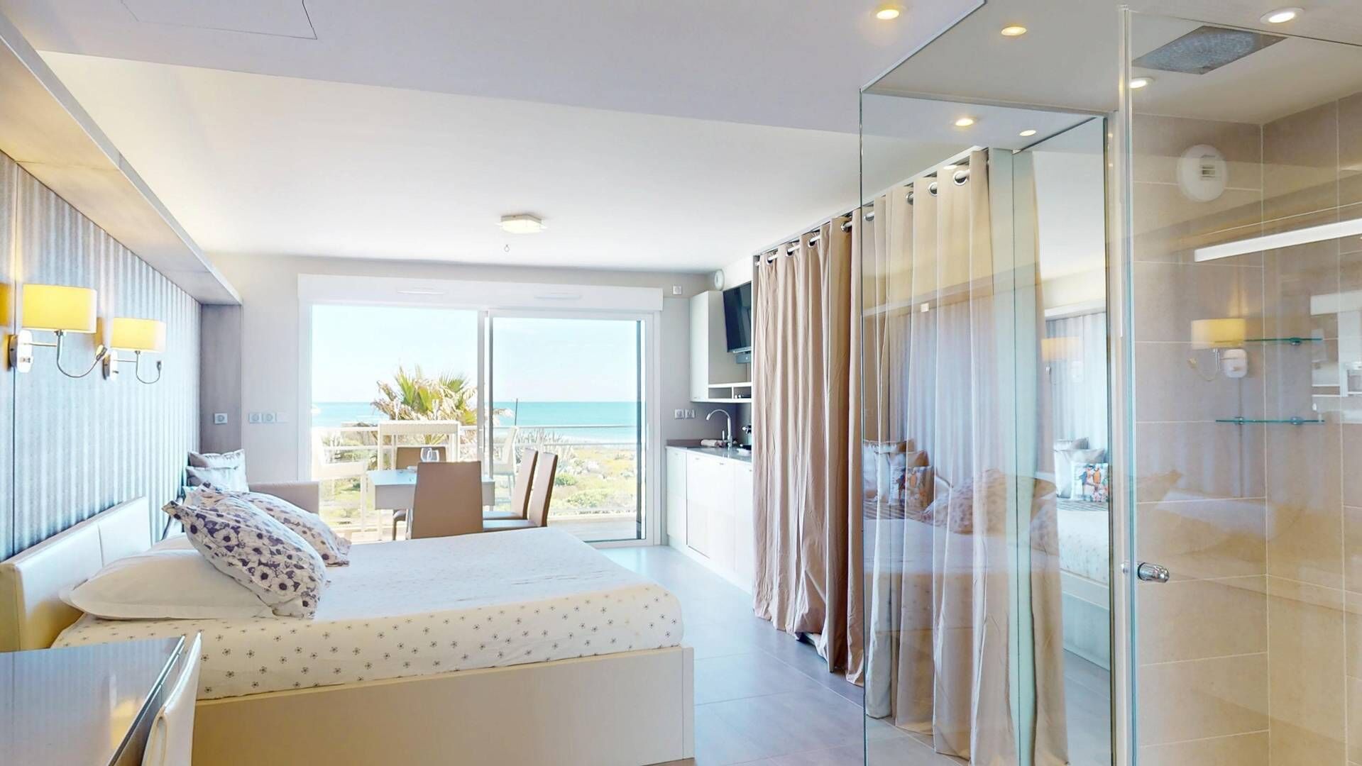 Una habitación de hotel moderna con una cama, un escritorio, un balcón con vista al mar y un box de ducha de vidrio.