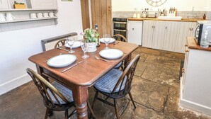 Comedor con mesa de madera dispuesta para cuatro personas, un reloj en la pared y una cocina al fondo.