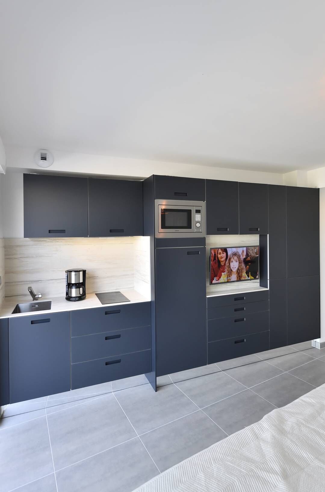 A modern kitchen with dark cabinetry, a built-in microwave, a coffee maker, and a TV screen displaying a video call.