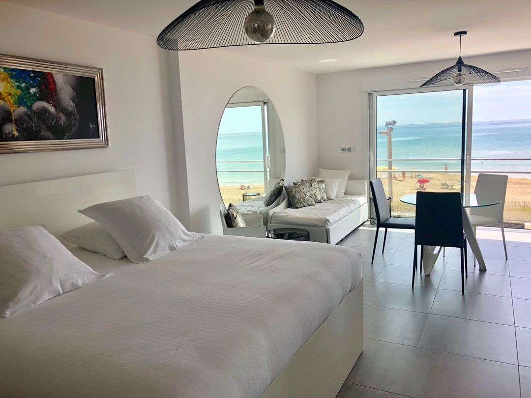 A modern hotel room with a large bed, a seating area with a view of the beach, and a dining table.