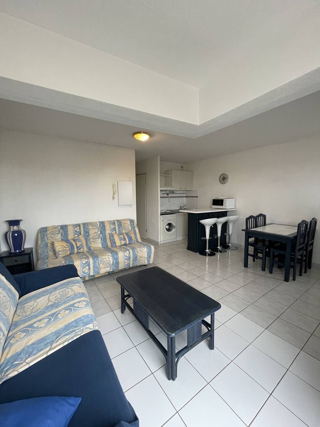 Ein Wohnzimmer mit einer Couch, einem Couchtisch, einem Esstisch mit Stühlen, einer Küchenzeile mit einer Mikrowelle und einer Waschmaschine.