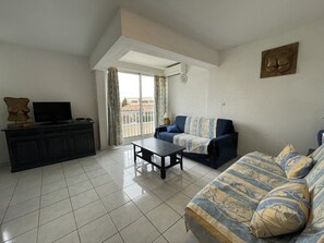Ein Wohnzimmer mit einer Couch, einem Couchtisch, einem Fernseher auf einem Schrank und einem Balkon mit Vorhängen.