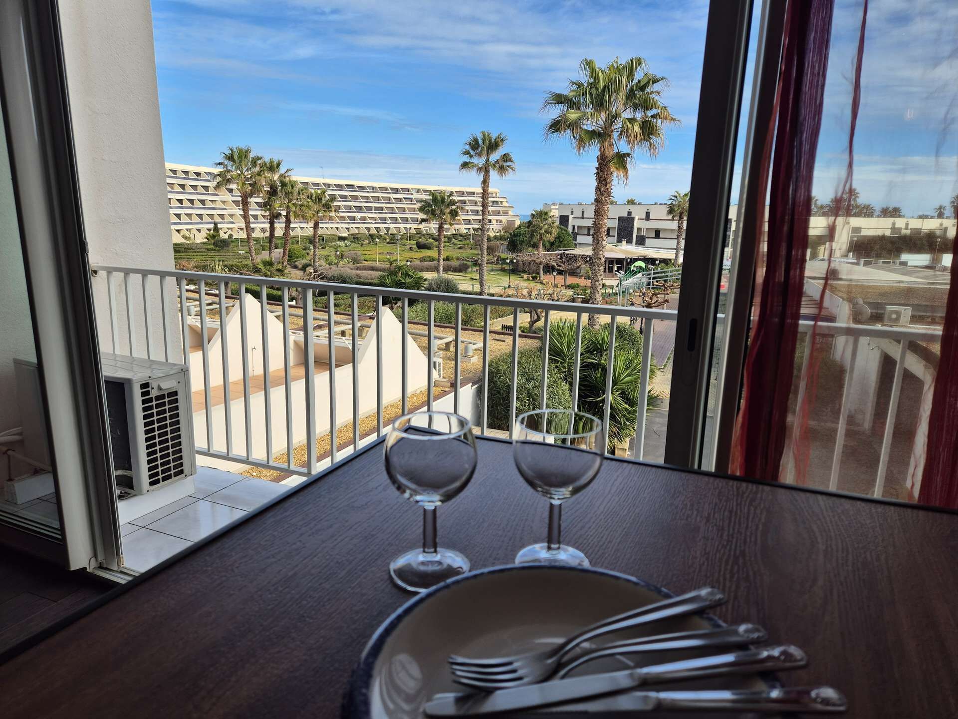 En balkong med utsikt över ett hotellkomplex, ett bord med vinglas och bestick samt ett fönster med gardiner.