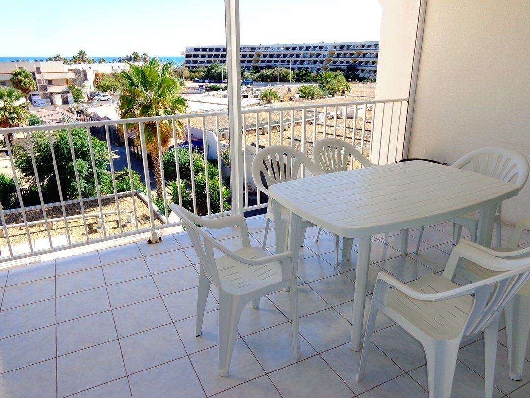 Un balcón con una mesa y sillas blancas, vista al mar y un edificio en el fondo.