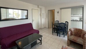 A living room with a sofa, a table, and a chair. There is a door leading to a kitchen area. - Natura Beach - Apartment NB303 - 8 people (AGDE)
