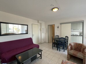 A living room with a sofa, a table, and a chair. There is a door leading to a kitchen area. - Natura Beach - Apartment NB303 - 8 people (AGDE)