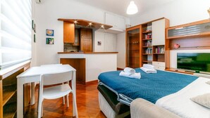 Een compacte kamer met een bed, een klein bureau, een stoel, een televisie en een kitchenette.