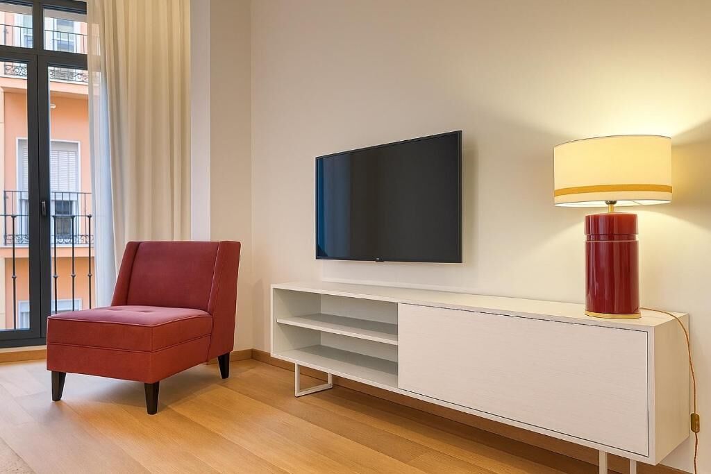 Un salon moderne avec un téléviseur à écran plat, un fauteuil rouge, un meuble de rangement blanc et une lampe.