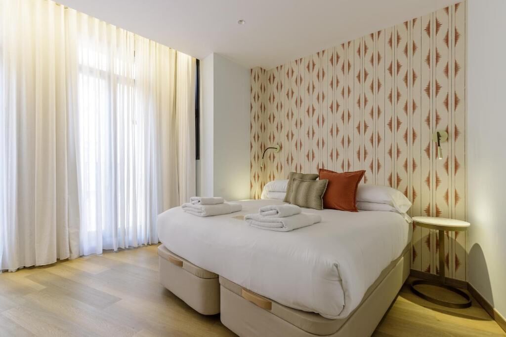 Une chambre d’hôtel avec un lit, des rideaux, un mur décoré de motifs, une table de chevet et une lampe.