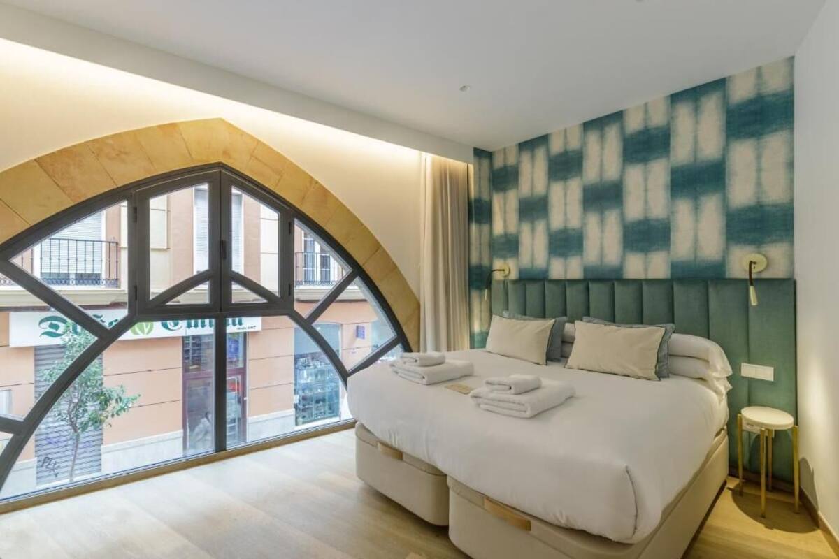 Habitació d'hotel amb una finestra gran d'arc, un llit amb roba blanca i un capçal de llit amb fons verd.