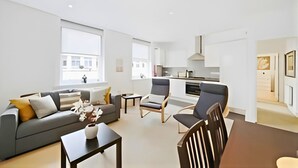 A modern living room with a sofa, chairs, a coffee table, and a kitchen area with cabinets and a stove. - Charming 1-Bed Apartment in Lewisham (London)