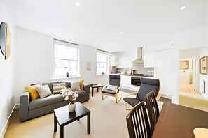 Ein modernes Wohnzimmer mit einer Couch, Sesseln, einem Couchtisch und einer Küche im Hintergrund.