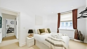 A modern bedroom with a bed, bedside table, lamp, and a window with curtains. - Charming 1-Bed Apartment in Lewisham (London)