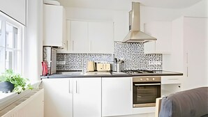 A modern kitchen with white cabinetry, a stainless steel range hood, and a black backsplash. - Charming 1-Bed Apartment in Lewisham (London)