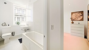 A modern bathroom with a bathtub, a sink, and a mirror. There is a window with a view of buildings outside. - Charming 1-Bed Apartment in Lewisham (London)