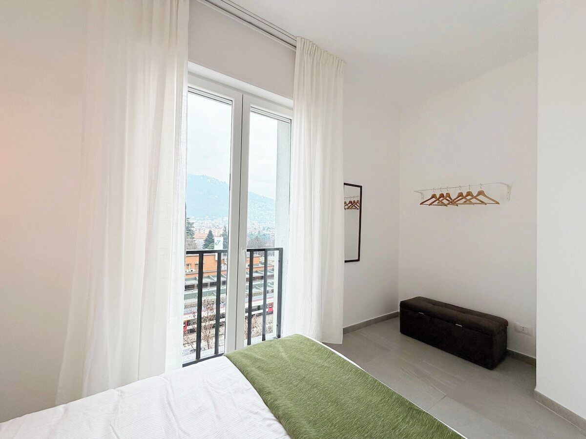 Una camera da letto con un letto, una panca, una finestra con tende e vista sui palazzi.