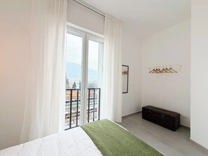 Una camera da letto con un letto, una panca, una finestra con tende e vista sui palazzi.
