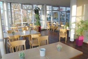 A spacious indoor area with multiple wooden tables and chairs, large windows, and potted plants.