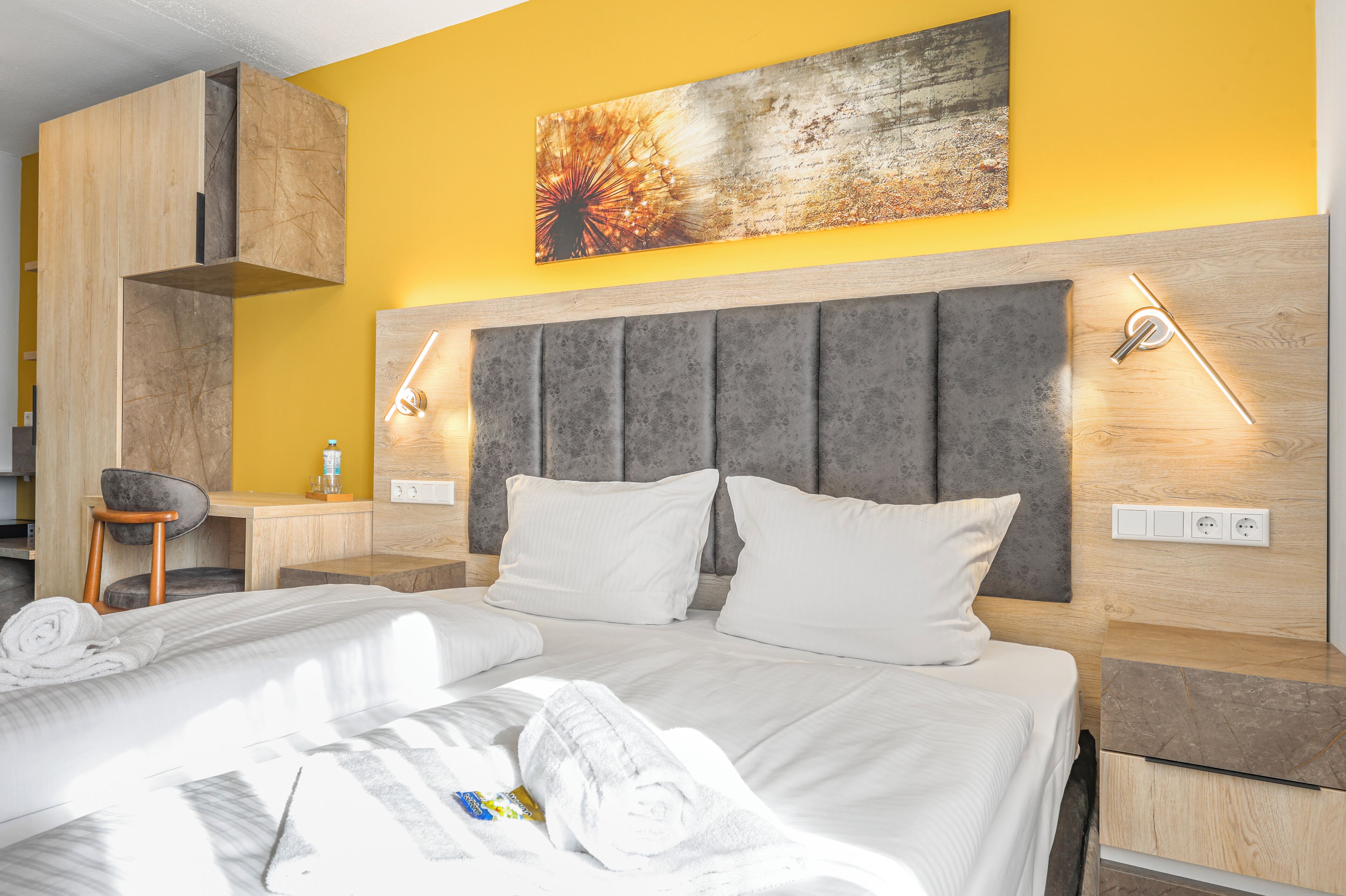 A hotel room with a bed, a desk, a chair, and a headboard with built-in lighting.