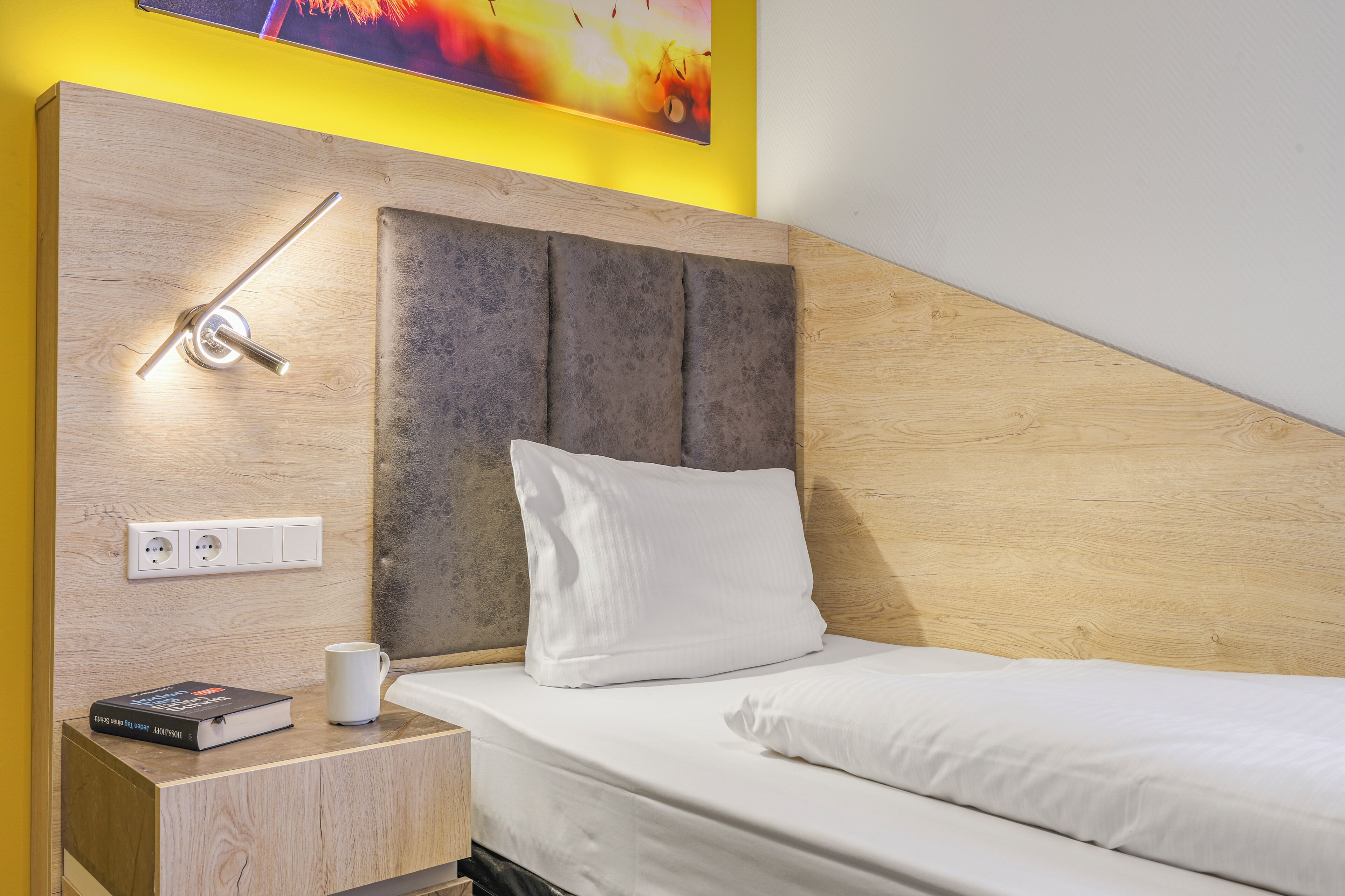 Una habitación de hotel moderna con cabecera de madera, una mesita de noche con un libro y una taza, y un enchufe.