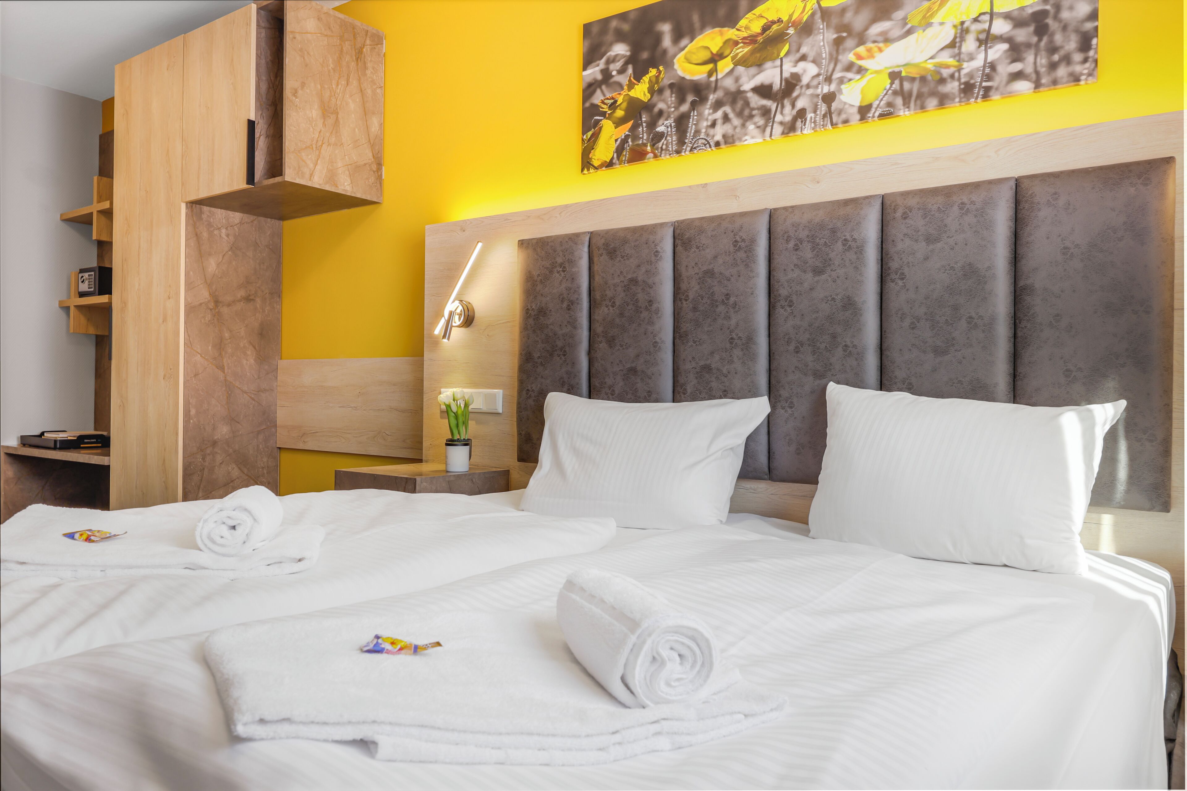 Habitación de hotel con una cama grande, un armario de madera, una mesita de noche con lámpara y una pared amarilla decorada con un cuadro floral.