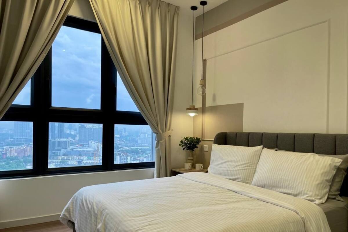 Ein Schlafzimmer mit großem Fenster, einem Bett, einem Nachttisch mit Lampe und Blick auf eine Stadtlandschaft.
