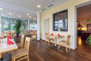 A spacious dining area with wooden tables and chairs, large windows, and a potted plant.