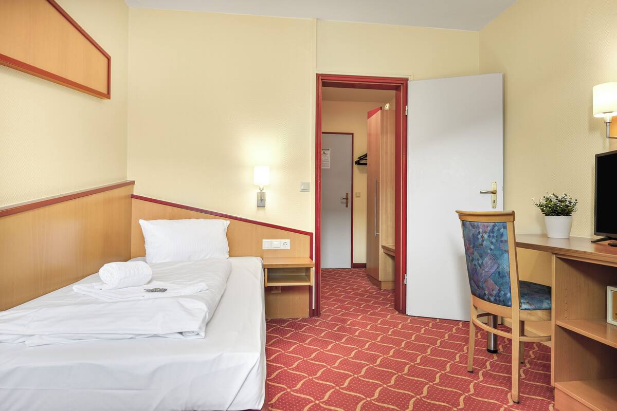 A hotel room with a bed, a desk, a chair, a TV, and a door leading to a hallway.
