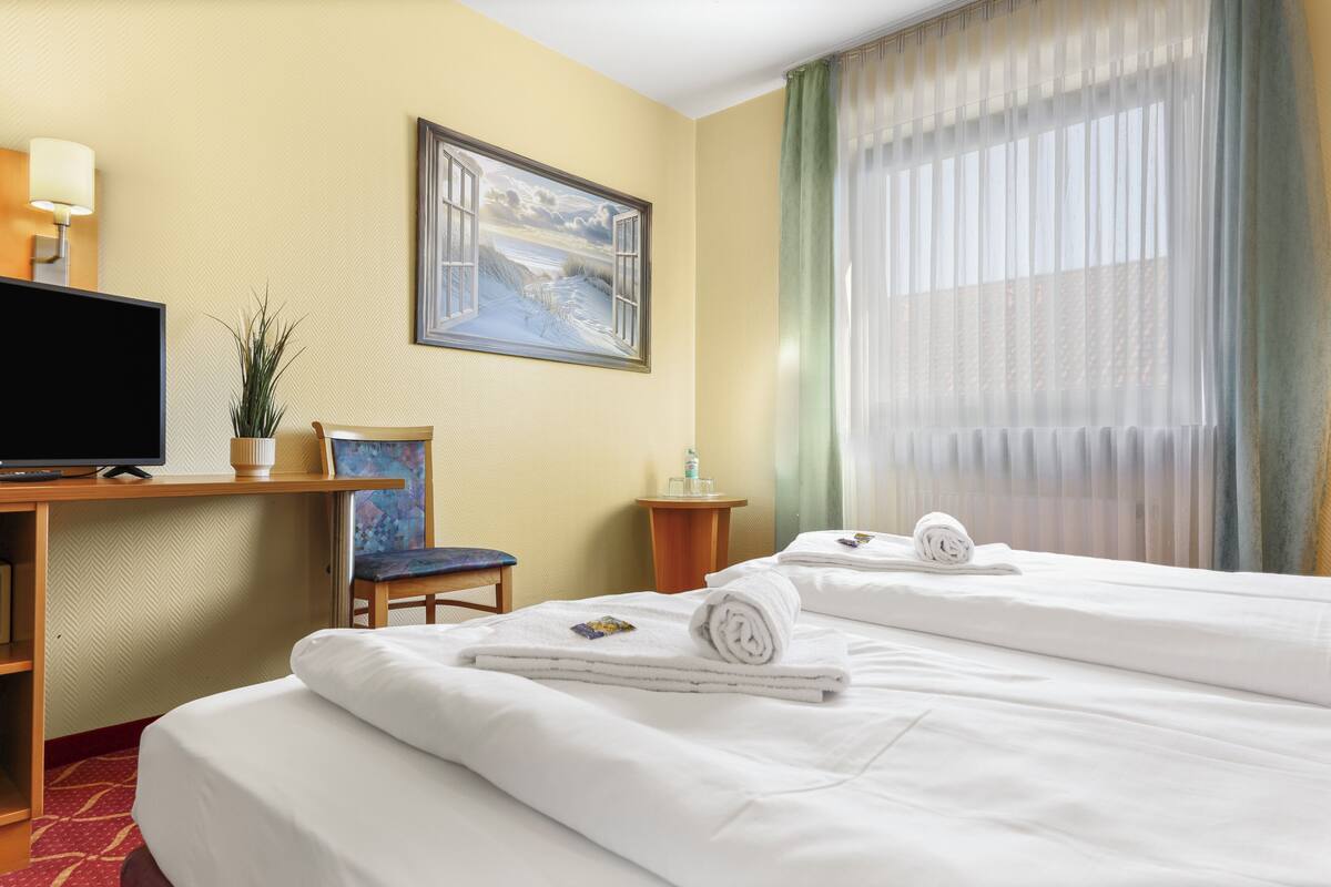 A hotel room with a bed, a desk with a TV, a chair, a painting, and a window with curtains.