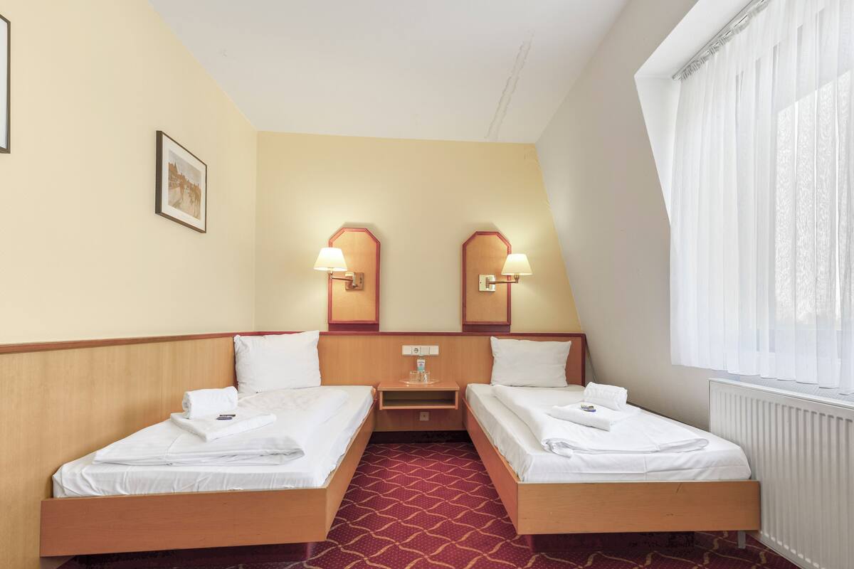 A hotel room with two single beds, a wooden headboard, a red patterned carpet, a small bedside table with a lamp, and a window with sheer curtains.