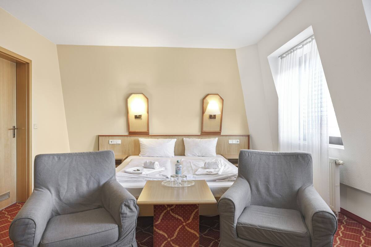 A hotel room with a bed, two armchairs, a small table, and a window with curtains.