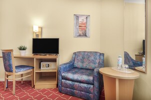 A hotel room with a TV, a patterned armchair, a small table with a glass and water bottle, a chair, and a wall-mounted picture.