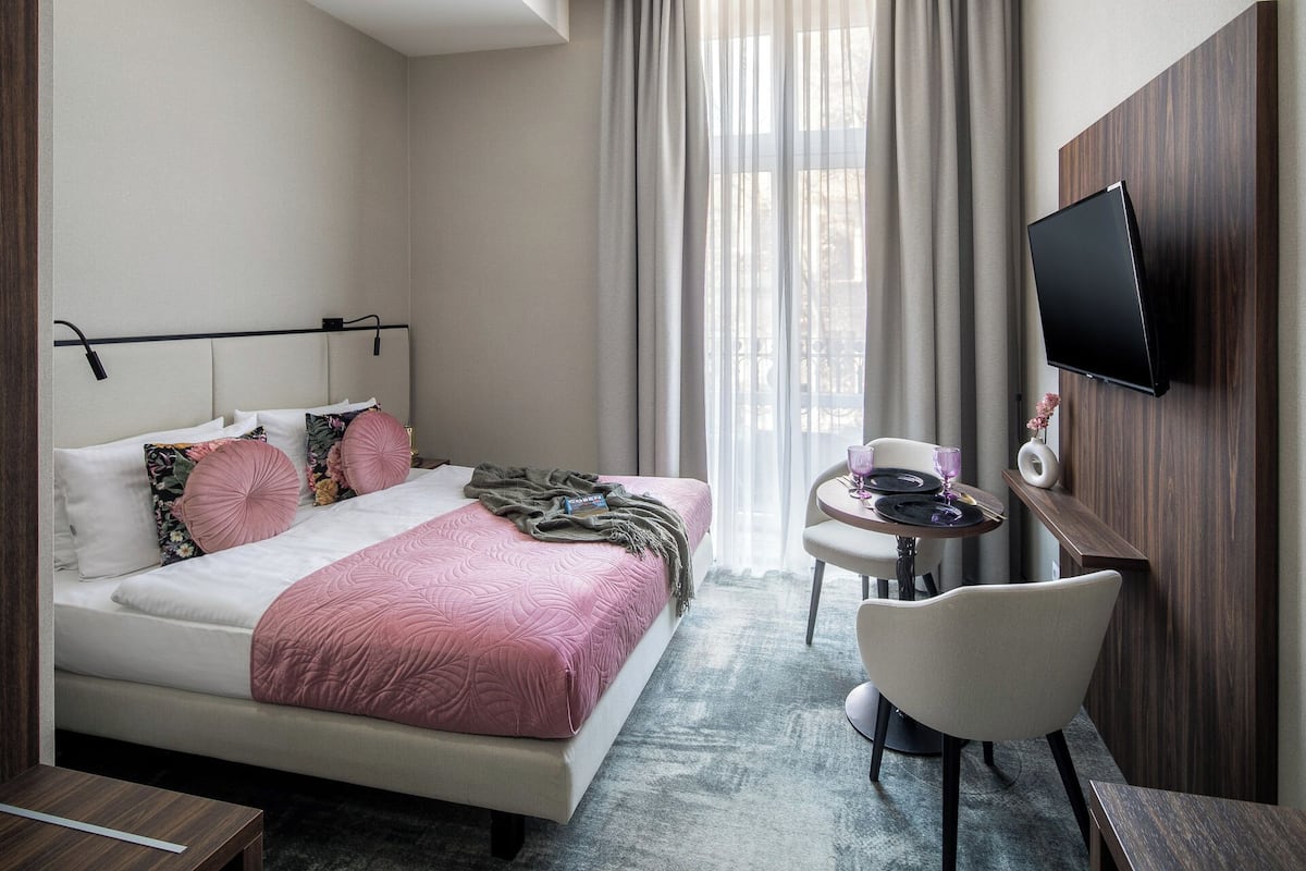 Quarto de hotel com cama, mesa de cabeceira, cadeira e televisão fixada na parede.