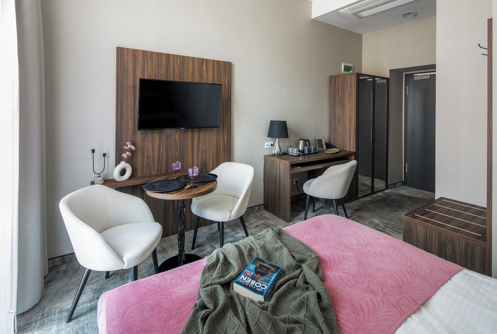 Et hotelværelse med en seng, to hvide stole, et skrivebord med tv, en lampe og et minibar.