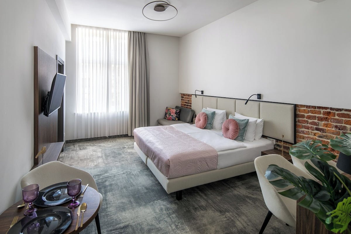 Quarto de hotel com cama, escrivaninha com cadeira, televisão e janela com cortinas.