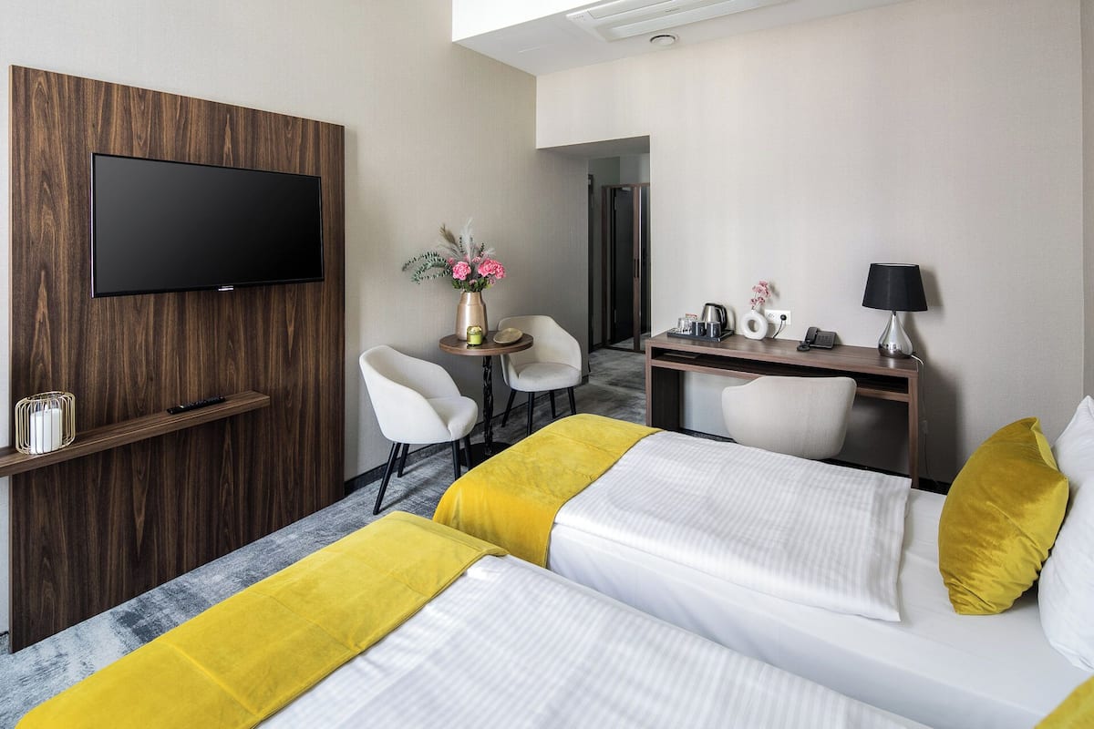 Quarto de hotel com televisão fixada num painel de madeira, duas camas com roupa de cama amarela e branca, uma secretária com candeeiro, uma cadeira e uma pequena mesa com um vaso de flores.