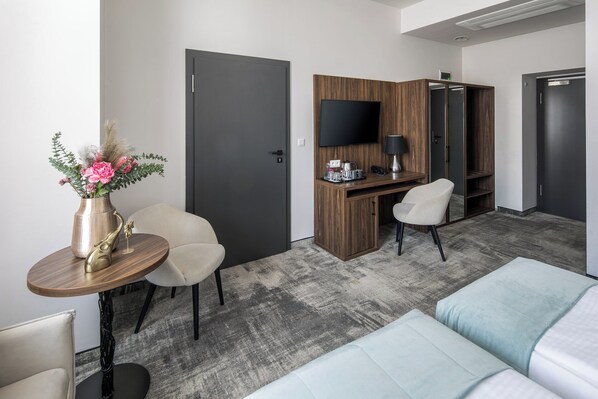 A hotel room with a bed, a desk with a computer, a chair, a small table with a vase of flowers, and a TV mounted on the wall.