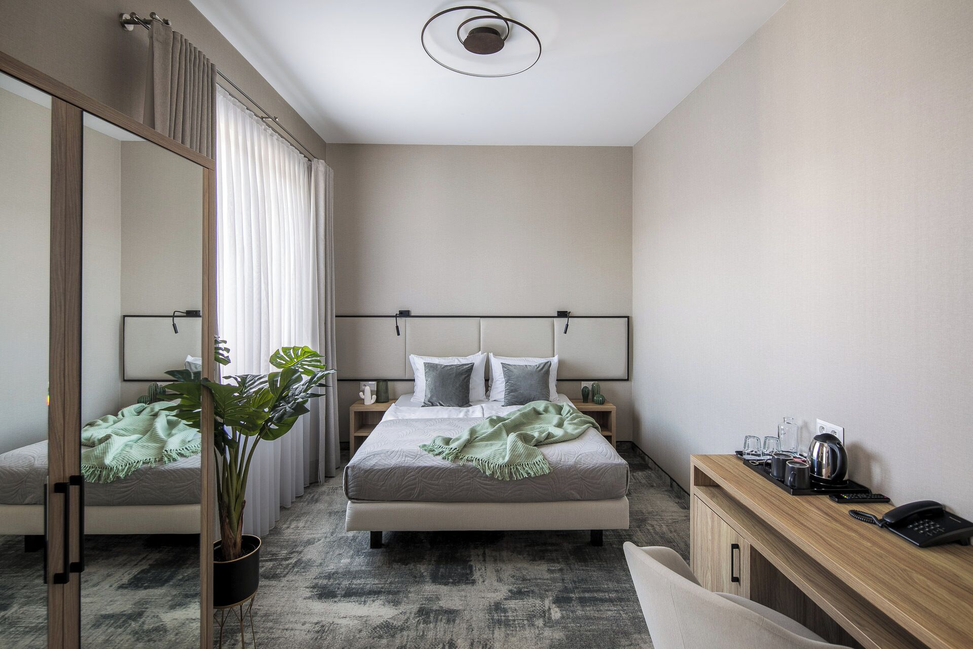 A modern hotel room with a large bed, bedside tables, a desk with a telephone, and a potted plant.