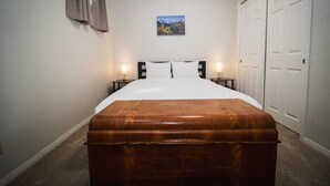 Un dormitorio con una cama, lámparas de noche, un cofre de madera, un cuadro en la pared y un armario.