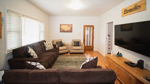 Una sala de estar con un sofá seccional marrón, dos sillones, un televisor de pantalla plana grande y un mueble de madera.