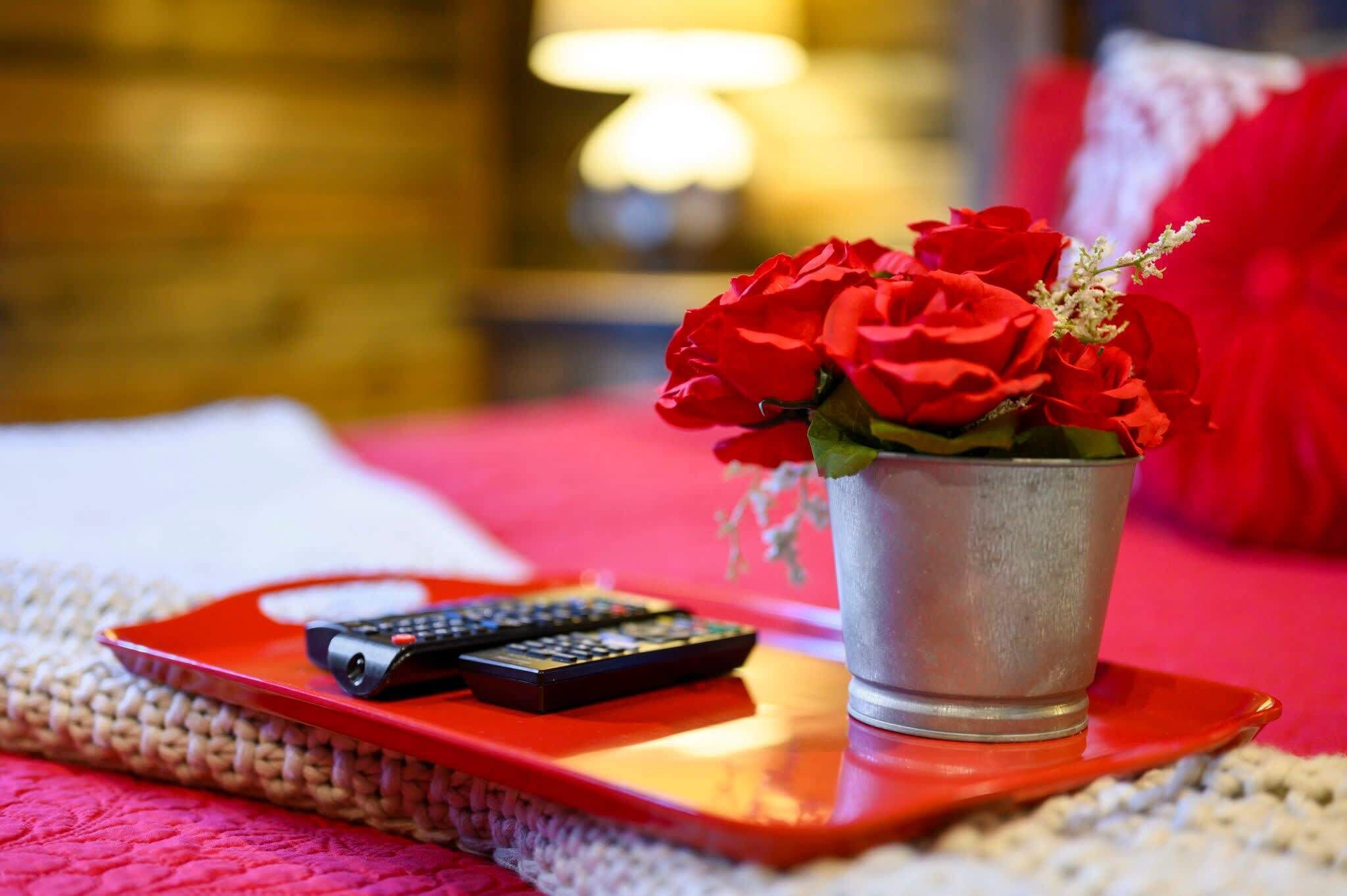 A bed with a red blanket, a metal bucket with red roses, and two remote controls on a red tray.
