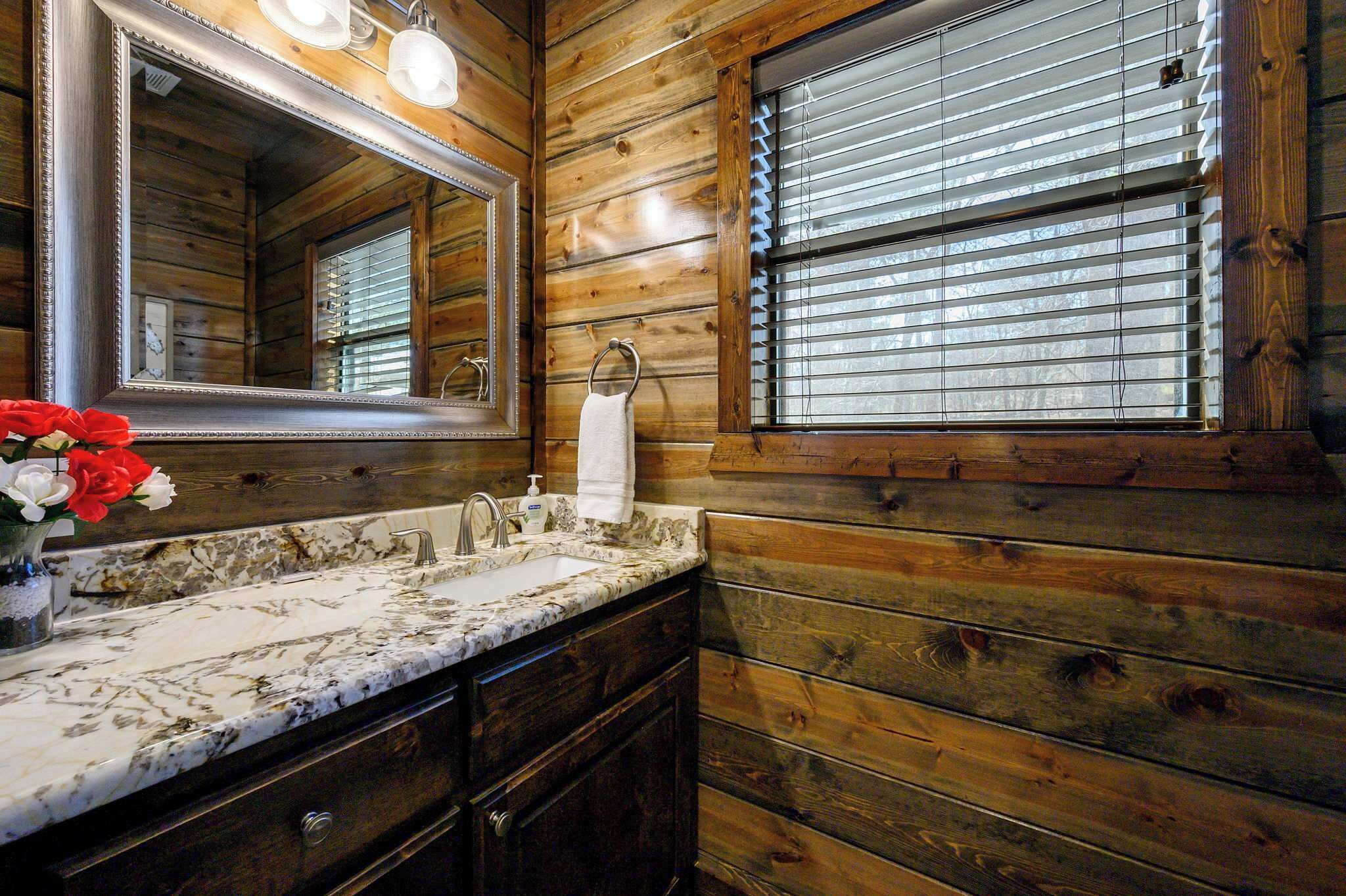 A wooden cabin-style bathroom with a marble sink, a large mirror, and a window with blinds.