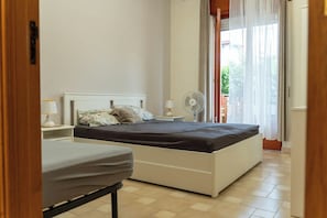 Een slaapkamer met een bed, nachtkastje, lamp en een staande ventilator.
