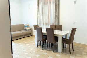 Een zithoek met een tafel en stoelen, een bank met een stap opgevouwen dekens, en een raam met gordijnen.