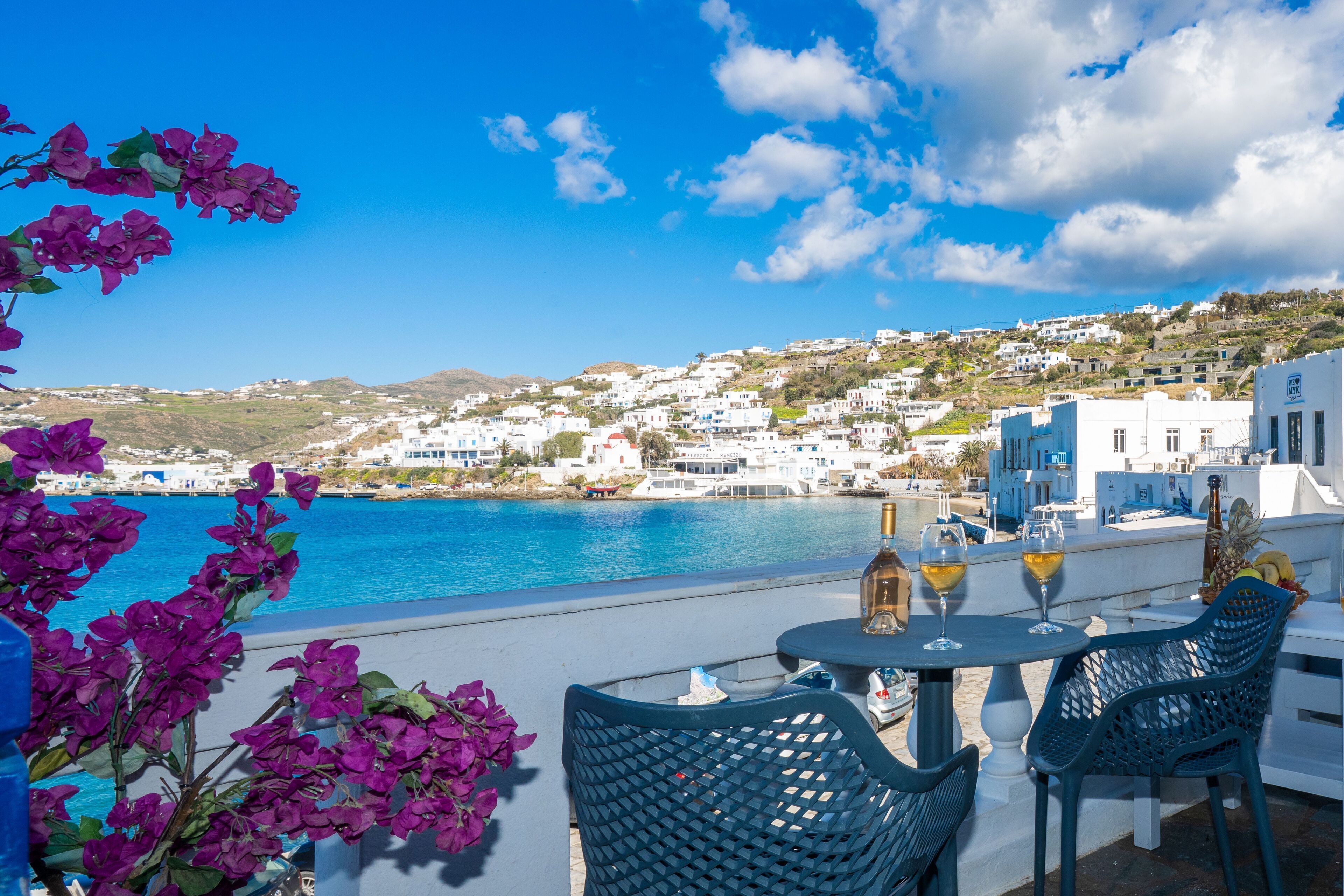 A seaside terrace with a view of white buildings, a table with chairs, and a bottle of wine.